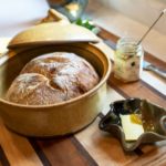 a horizontally framed photograph shows a relatively close up view of a clay coyote bread baker, a small glass jar of honey, a clay coyote little dipper and a small block of butter with a knife. all of this is resting on a wooden cutting board. the bread baker is glazed in yellow salt, with the lid resting on the "back" (relative to the viewpoint) circular lip of the bread bakers base. the bread baker has been used to bake a fresh loaf of bread, which is still resting inside the bread bakers bottom. the bread has a nice crust developed with light and dark brown coloration and a light dusting of white flour still remains on sections of the crust. to the lower right of the bread baker base is a clay coyote little dipper serving dish. it is 6 pointed. inside the little dipper is a piece of butter, with honey on its upper right corner. there is more honey resting to the right of the butter inside of the small dish. to the right of the bread baker, and "behind" the little dipper is a small 6 sided glass jar of honey. the label is white, with black writing and the words "raw local honey" can be made out, despite being out of focus. there is a dinner spoon resting inside the jar, the jars lid is off and unseen else where. to the right of the small honey jar is a kitchen butter knife. only the handle is fully visible, while the thinner blade is partially obscured by the honey jar. partially behind to the upper left of the bread baker is a small tray of butter. the knob of butter in the little dipper is clearly cut from this larger half pound(?) piece of butter. a silver butter knife handle is visible resting next to the butter. the wooden cutting board everything is resting on has wide strips of light and medium colored wood, with thin dark strips of dark wood dividing each wide piece from the next.