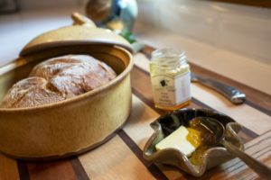 a horizontally framed photograph shows a close up of a clay coyote bread baker, glazed in yellow salt, and a clay coyote little dipper serving dish. the bread baker has been used to bake a fresh loaf of bread, which is still resting inside the bread bakers base. the lid to the bread baker is resting on the "back" (relative to the viewpoint) of the circular rim. the handle on the top of the bread baker lid is visible. to the right of the bread baker is a small glass jar with flat sides, it appears to be 6 sided. the jar has its lid off, and is a jar of honey. the jar has a white label with black writing. the words "raw LOCAL Honey" can be made out (the jar is slightly out of focus, and the rest of the writing is too small to identify). partially behind the small honey jar, and partially to the right of it is a silver colored butter knife. only the thicker handle is totally visible, while the bladed portion of it is mostly obscured by the honey jar. in front of the honey jar (and to the lower right of the bread baker) is a clay coyote little dipper serving piece. it is the circular 6 pointed style. it is glazed in mint chip. the little dipper has a tablespoon or so of butter resting inside of it. there is also a silver colored dinner spoon resting inside of it also. the spoon has been used to scoop up some honey into the little dipper. the honey is spilling onto the butter knobs lower right corner, and some onto the little dipper itself, and there is still a good amount still on the spoon head itself. the little dipper, the honey, the butter knife and the bread baker are all resting on a wooden cutting board. the wooden cutting board has different stripes of dark and light colored wood. each of these wider stripes is divided from the next by a thin dark colored stripe of wood. out of focus in the background is a white kitchen countertop. resting on the countertop, behind the wooden cutting board with the bread baker and all is a clay coyote vase glazed in joes blue. it is partially cut off the the top boarder of the photograph, and is out of focus.