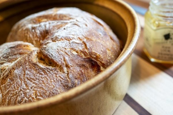 a horizontally framed photograph shows a very close view of the base of a clay coyote bread baker. the bread baker is glazed in yellow salt, giving it a yellow coloring with little flecks of darker yellow or a very very light brown. the bread baker has been used to bake a fresh loaf of bread. the loaf of bread is whole and unsliced. slightly out of frame on the right side of the photograph is a out of focus small jar of honey, with the lid off. the jar is glass with a white label with black writing. the photograph was taken close enough that only about 75% of the bread baker is visible. the bread baker and the jar of honey are resting on a wooden cutting board. the wooden cutting board has wide strips of light and dark colored wood, with thin dark colored strips dividing the wide ones.