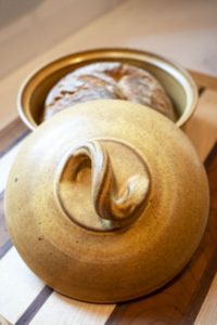 a vertically framed photograph shows a close up view of a clay coyote bread baker glazed in yellow salt. the lid is resting on the circular lip of the base, and an angle that allows the viewer a "top down" view of the bread bakers lid. the lid handle is shaped like a reverse "S" with the handle itself being twisted to provide a visually pleasing aesthetic. the base of the bread baker has a whole loaf of freshly baked bread inside, that is visible behind the lid, relative to the viewer. the bread loaf if whole and unsliced. the bread baker is resting on a wooden cutting board. the cutting board has large strips of light and dark colored wood, which is divided by thin stripes of dark colored wood. the bread baker and wooden cutting board are sitting on a white kitchen counter top. the photograph is lit by both warm yellow light and white natural light.