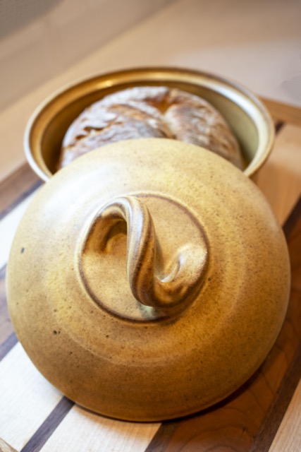 a vertically framed photograph shows a close up view of a clay coyote bread baker glazed in yellow salt. the lid is resting on the circular lip of the base, and an angle that allows the viewer a "top down" view of the bread bakers lid. the lid handle is shaped like a reverse "S" with the handle itself being twisted to provide a visually pleasing aesthetic. the base of the bread baker has a whole loaf of freshly baked bread inside, that is visible behind the lid, relative to the viewer. the bread loaf if whole and unsliced. the bread baker is resting on a wooden cutting board. the cutting board has large strips of light and dark colored wood, which is divided by thin stripes of dark colored wood. the bread baker and wooden cutting board are sitting on a white kitchen counter top. the photograph is lit by both warm yellow light and white natural light.