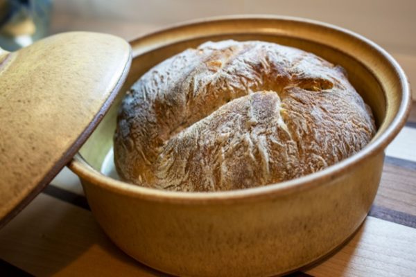 a horizontally framed photograph shows a close up view of a clay coyote bread baker glazed in yellow salt. the bread baker has just been used to bake a fresh loaf of bread. the bread loaf is sitting in the bread bakers base, with the bread baker cover resting on the left side of the circular lip, going partially out of frame (on the left edge of the photo). the bread loaf is whole and unsliced. the bread baker is resting on a multicolored wooden cutting board. the photograph is lit by a mixture of white and warm yellow lighting.