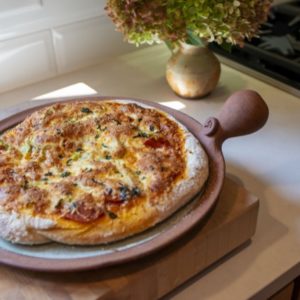 a horizontally framed photograph shows a full view of a clay coyote pizza stone that has been used to bake a fresh pizza. the pizza is almost completely covering the pizza stones cooking area. the cooking area of the pizza stone is glazed in coyote grey to denote that it is flameware. the pizza stones rim/edge, secondary handles and primary bulbous handle are all unglazed and show off the natural reddish brown of the clay. the pizza stone is sitting on a square wooden butchers block style cutting board (it appears to be over an inch thick of wood). the pizza stone is resting on the cutting board and the cutting board is resting on a white kitchen countertop. behind the pizza/butchers block (relative to the view point) is a clay coyote small vase glazed in feather with a large bouquet of flowers resting in it. further "back" is a gas stovetop range, with only one burner visible.