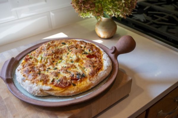 a horizontally framed photograph shows a full view of a clay coyote pizza stone that has been used to bake a fresh pizza. the pizza is almost completely covering the pizza stones cooking area. the cooking area of the pizza stone is glazed in coyote grey to denote that it is flameware. the pizza stones rim/edge, secondary handles and primary bulbous handle are all unglazed and show off the natural reddish brown of the clay. the pizza stone is sitting on a square wooden butchers block style cutting board (it appears to be over an inch thick of wood). the pizza stone is resting on the cutting board and the cutting board is resting on a white kitchen countertop. behind the pizza/butchers block (relative to the view point) is a clay coyote small vase glazed in feather with a large bouquet of flowers resting in it. further "back" is a gas stovetop range, with only one burner visible.