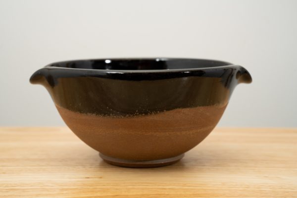 the photograph shows a clay coyote cassoulet serving bowl glazed in midnight black resting on a light colored wooden surface. the bowls outside bottom is unglazed, and shows the natural color of the clay. the inside and outside upper lip are glazed in midnight black. the bowl is resting flat on the wooden surface. the background is a white wall.