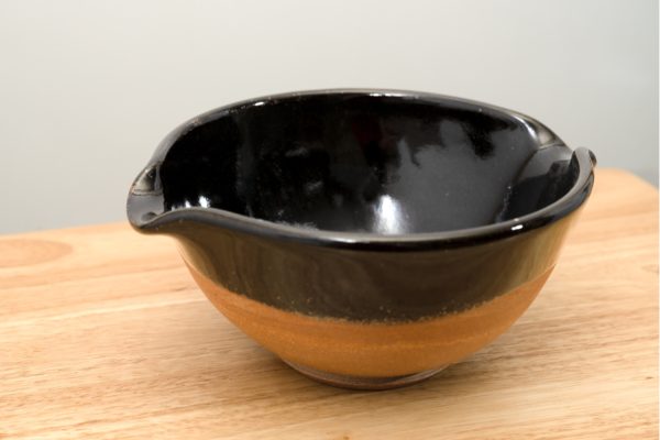 the photograph shows a low angle view of a clay coyote cassoulet serving bowl glazed in midnight black. the bowl is resting on a small light colored wooden table. the bowl is glazed in midnight black on the inside and halfway down the outside of the bowl. the unglazed portion on the outside bottom of the bowl is reddish brown, the natural color of the clay. the background is a plain white wall. the photograph is well lit with white light.