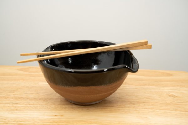 the photograph shows a clay coyote cassoulet bowl glazed in midnight black with a pair of chopsticks resting on top of the bowl. the bowl is glazed black on the inside and the upper half of the outside. the bottom outside of the bowl is unglazed, and shows the natural color of the clay. the bowl is resting flat on a light colored wooden surface. the background is a plain white wall. the photograph is well lit with white light.