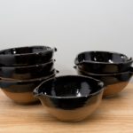 the photograph shows 6 cassoulet bowls glazed in midnight black resting on a small light colored wooden table. there is a bowl by itself in the front, then to the back left is a stack of three bowls. finally to the back right there is a stack of two cassoulet bowls. all the bowls are glazed on the inside and the upper outside lip in midnight black. the outside bottoms of all the bowls are unglazed and show the natural reddish brown clay color. the background is a plain white wall and the photograph is well lit with white light.