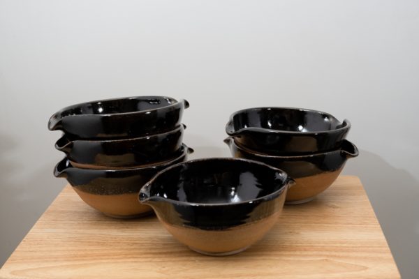 the photograph shows 6 cassoulet bowls glazed in midnight black resting on a small light colored wooden table. there is a bowl by itself in the front, then to the back left is a stack of three bowls. finally to the back right there is a stack of two cassoulet bowls. all the bowls are glazed on the inside and the upper outside lip in midnight black. the outside bottoms of all the bowls are unglazed and show the natural reddish brown clay color. the background is a plain white wall and the photograph is well lit with white light.