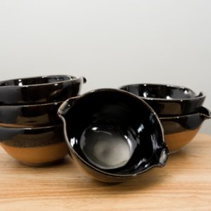 the photograph shows 6 cassoulet bowls glazed in midnight black resting on a small light colored wooden table. there is a bowl by itself in the front, which is tilted to show the inside of the bowl, and how the inside is completely glazed in black. to the back left is a stack of three bowls. finally to the back right there is a stack of two cassoulet bowls. all the bowls are glazed on the inside and the upper outside lip in midnight black. the outside bottoms of all the bowls are unglazed and show the natural reddish brown clay color. the background is a plain white wall and the photograph is well lit with white light.