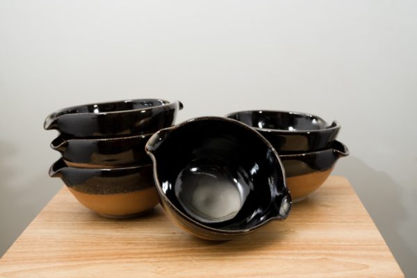 the photograph shows 6 cassoulet bowls glazed in midnight black resting on a small light colored wooden table. there is a bowl by itself in the front, which is tilted to show the inside of the bowl, and how the inside is completely glazed in black. to the back left is a stack of three bowls. finally to the back right there is a stack of two cassoulet bowls. all the bowls are glazed on the inside and the upper outside lip in midnight black. the outside bottoms of all the bowls are unglazed and show the natural reddish brown clay color. the background is a plain white wall and the photograph is well lit with white light.