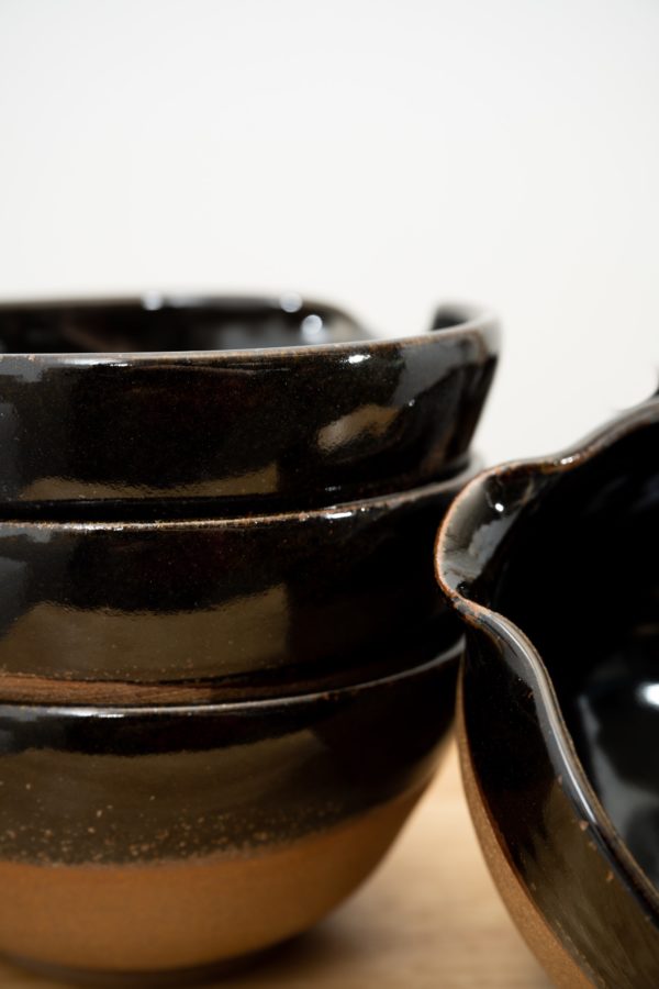 the photograph shows 3 clay coyote cassoulet bowls glazed in midnight. the photograph is very close up, none of the three cassoulet bowls are fully in frame. there is two bowls stacked on top of each other on the left. on the right is the pour spout of one cassoulet bowl tilted on its side. only the area immediately around the left pour spout is visible in the photograph. in the upper right corner there is a little bit of white background visible, it is out of focus. the photograph is lit with white light.