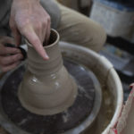 a horizontally framed photograph shows a clay coyote yunomi being formed on a pottery wheel. the potter is seated (mostly out of frame) to the upper left with basically only his hands and knee visible. he is in process of making a clay coyote yunomi, forming it out of a larger lump of clay. he is holding a shaping tool to add grooves or ridges to the sides of the cup, with his right hand, and his left hand is being used as a stabilizer and guide