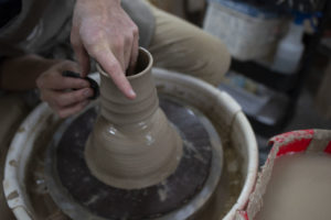 a horizontally framed photograph shows a clay coyote yunomi being formed on a pottery wheel. the potter is seated (mostly out of frame) to the upper left with basically only his hands and knee visible. he is in process of making a clay coyote yunomi, forming it out of a larger lump of clay. he is holding a shaping tool to add grooves or ridges to the sides of the cup, with his right hand, and his left hand is being used as a stabilizer and guide