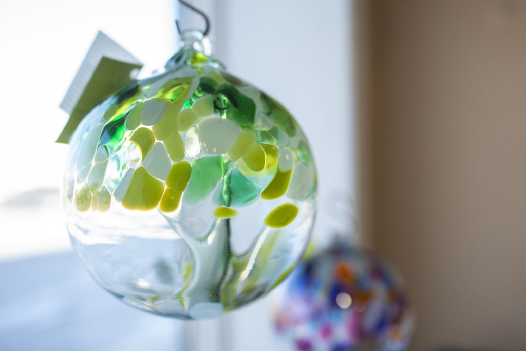 a horizontally framed photograph shows a kitras blown glass ball, hanging in a window. the blown glass sphere has been blown to resemble a tree on the inside, made of clear glass, the bottom of the sphere is clear also. the top half of the orb is multicolored with greens and whites representing the leaves of the "tree" contained within. the background is out of focus.