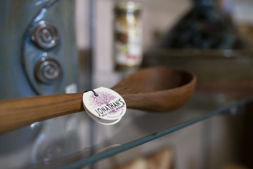 a horizontally framed photograph shows a very close picture of a Jonathan's wild cherry spoon, sitting on a glass shelf, behind it and to the left part of a clay coyote wine chiller/utensil holder is visible.
