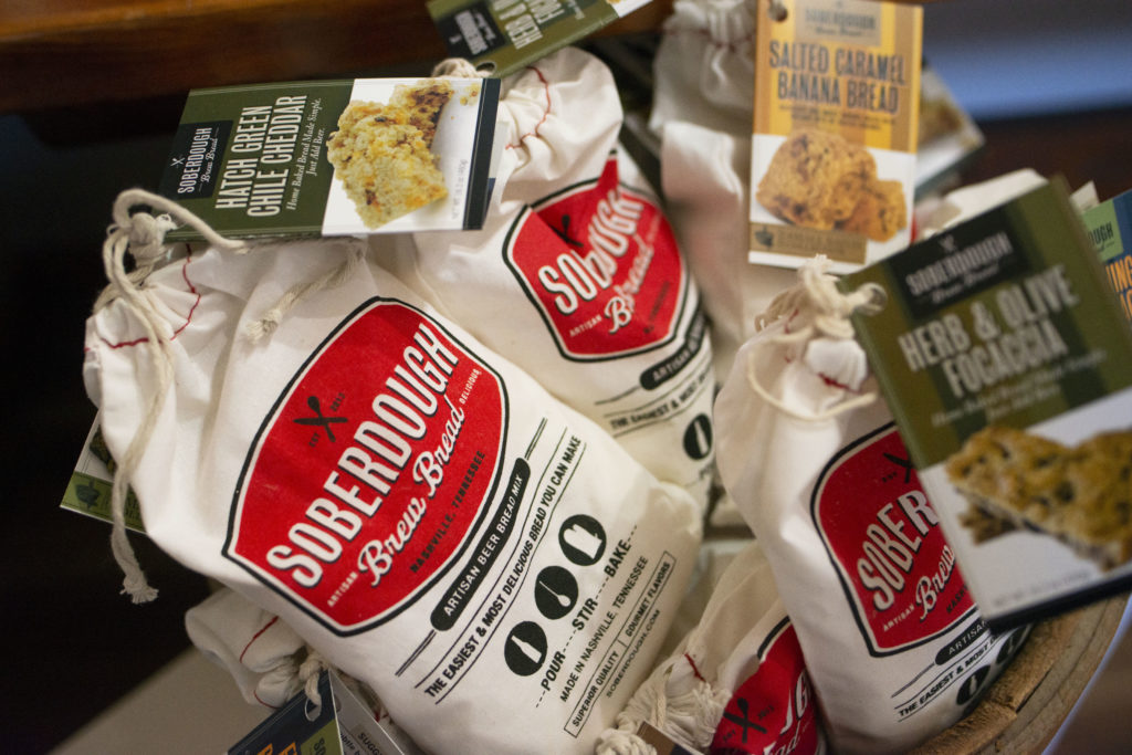 a horizontally framed photograph shows a close up photograph of Soberdough brand mixes of bread. there is a tag visible for herb and olive focaccia and hatch green chile cheddar. they are white cloth bags with red badge branding of their logo.