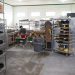 a horizontally framed photograph shows the clay coyote shop floor during the work day. sunlight is streaming through the four windows on the back wall. the left edge of the photograph shows a 6 shelf wire rack with pottery arranged on it. almost by the back wall a potter is working on slab pottery, in the mid ground of the photograph just left of center a potter in a plaid shirt and khakis is forming a yunomi cup "off the hump" (forming a cup out of the top of a larger amount of clay). closer to the right side of the photograph a potter is partially hidden by a cart filled with pottery, and covered by plastic sheets. the potter is wearing a black shirt and blue jeans.