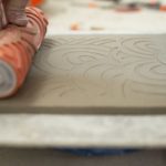a horizontally framed photograph shows the process of how the pattern on the bottom side of a baking dish is imprinted into the clay. the potter, (only parts of the fingertips are in the photograph) is holding a roller, with a design cut into it, and has just rolled it across the surface of the flat clay laying on her work surface. the photograph is a very close up, with only the slab, roller and the immediate work surface around the slab visible.