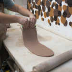 a horizontally framed photograph shows a potter doing slab work with clay. only the potters torso is visible (on the left edge of the photograph) and her forearms/ hands. she has just rolled out a long thin piece of raw clay, and is lifting half of this piece up off the work surface. the work surface is white, and there is a white wall behind it visible, with various painted dots (part of a larger un-pictured mural on the the wall). there is a rolling pin visible on the work surface. the potter is wearing a apron.