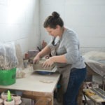 a horizontally framed photograph shows a clay coyote potter in process of making a baking dish. she is standing at a table, near the corner of the room. the bottom of the baking dish is visible on a work surface, and she is in process of attaching the wall of the piece. she is looking down at the piece. the room is lit with sunlight. she has a grey shirt and blue jeans on, with a clay colored apron on. around her are various tools she uses.
