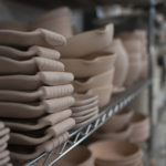 a horizontally framed photograph shows a close up of clay coyote pottery that has been bisque fired, and is stacked on wire racking, awaiting glazing. the lights are off in the shop. the closest pottery is on the left and moving to the right they are further and further away from the viewers perspective. the closest pottery is small trays without handles, then a stack of 9 large trays with handles, then another stack of small trays without handles. after that is 4 small skillets, and an ern. after that the pottery is too out of focus.