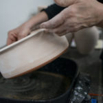 a horizontally framed photograph shows a close up shot of a clay coyote bread baker bottom. the bread baker has been bisque fired, and is in process of being glazed. the artist's hands are coming from out of frame on the top of the photo. the background out of focus.