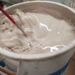 a horizontally framed photograph shows a close up of the top of a clay coyote glazing bucket. the surface of the glaze is turbulent, as a red mixing attachment is currently mixing the glaze. the photograph is close, close enough that not all of the top of the bucket is visible. the upper middle of the buckets rim is cut out of frame, with the rest of the rim being visible. the bucket is white.