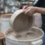 a horizontally framed photograph shows the potter pouring glaze out of a clay coyote bread baker bottom, having just submerged it in the glaze and is in process of pouring it out of the pottery. the entire piece is coated in the glaze. the potter is standing partially out of frame on the right side of the photograph, they are wearing a black long sleeve shirt and jeans, with the sleeves pulled up to mid-forearm. the background is out of focus, but shows some shelving, and another bucket of glaze, with a redder glaze visible around the rim of it.