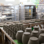 a horizontally framed photograph shows a behind the scenes shot of clay coyote pottery drying. the lights in the shop are not on, and a stream of bright morning light is visible in the upper left corner of the photograph. there are empty carts with plastic sheets on them in the background. in the fore ground is (starting nearest to the viewer) vases, then around 30 yunomis, then on the next table 14 dinner plates.