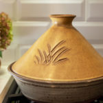 a horizontally framed photograph shows a clay coyote tagine in moroccan sand glaze (dark yellow with brown). The bottom of the tagine is clay coyote flameware, and glazed in coyote grey (grey with dark speckling through out). the tagine is sitting on the leftmost burner on a kitchen range. further left is a white kitchen countertop, with a clay coyote small vase in feather filled with a flower bouquet. the angle of the photograph is low, lower then eye level, but the top of the tagine is still visible.