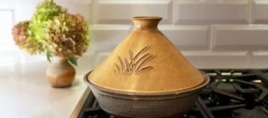 a horizontally framed photograph shows a clay coyote tagine in moroccan sand glaze (dark yellow with brown). The bottom of the tagine is clay coyote flameware, and glazed in coyote grey (grey with dark speckling through out). the tagine is sitting on the leftmost burner on a kitchen range. further left is a white kitchen countertop, with a clay coyote small vase in feather filled with a flower bouquet. the angle of the photograph is low, lower then eye level, but the top of the tagine is still visible.