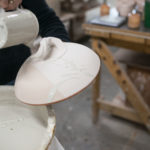 a horizontally framed photograph shows the glazing process of a clay coyote bread bakers lid. the lid is is being held from the bottom, with the handle side pointing up, the way it would be resting on a shelf/on the bread baker bottom. the lid has been dipped into 2 different glazes (all the glazes at this point are shades of grey, one more reddish, one darker, one lighter. the potter is using her other hand to drizzle glaze in artistic splashes over the top of the lid with the third glaze. the potter is using a small, but deep cylindrical measuring cup to pour the glaze over. the measuring cup is completely coated in the glaze, it must have been just dipped completely into the glaze. the right of the photograph is out of focus and shows the edge of the work space behind the potter. the only visible part of the potter in the photograph is part of her black shirt.