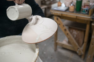 a horizontally framed photograph shows the glazing process of a clay coyote bread bakers lid. the lid is is being held from the bottom, with the handle side pointing up, the way it would be resting on a shelf/on the bread baker bottom. the lid has been dipped into 2 different glazes (all the glazes at this point are shades of grey, one more reddish, one darker, one lighter. the potter is using her other hand to drizzle glaze in artistic splashes over the top of the lid with the third glaze. the potter is using a small, but deep cylindrical measuring cup to pour the glaze over. the measuring cup is completely coated in the glaze, it must have been just dipped completely into the glaze. the right of the photograph is out of focus and shows the edge of the work space behind the potter. the only visible part of the potter in the photograph is part of her black shirt.