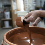 a horizontally framed photograph shows the process of glazing a clay coyote yunomi wine cup. the potter is standing on the right side of the photograph, with the glaze bucket taking up the lower right portion of the photograph. the potter is using one hand to hold the yunomi at an angle, pouring out the excess glaze from the inside. the outside of the yunomi shows it has just been dipped into the brown colored glaze. the area directly around the potters fingers remain unglazed. the potter is wearing a black long sleeve t-shirt with the sleeves pulled up to mid forearm. the background is out of focus but shows the workspace.