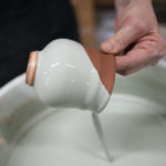 a horizontally framed picture shows a clay coyote yunomi wine cup in the process of being glazed. the potters hand is visible coming from the upper mid right of the frame, they are holding the lip edge of the yunomi in a pinch grip with thumb and pointer finger. the yunomi has just been dipped into the glaze, and is angled with the mouth pointed slightly down, with glaze pouring out of the inside, back into the bucket.