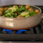 a horizontally framed photograph shows a close up of a clay coyote flameware cazuela filled with cooking veggies and small potatoes. the cazuela is sitting on a lit gas burner, with blue flame visible underneath. the cazuela is glazed in coyote grey (grey with dark speckling through out). the cazuelas main handle is pointed to the right. the vegetables within include: small whole yellow skin on potatoes, broccoli florets, halved brussel sprouts, whole green pea pods and some slices of red bell pepper. course black pepper has been sprinkled onto the cooking vegetables, and they all have a light sheen to them. to the left of the stove top gas range, is a white kitchen countertop. on the countertop is a clay coyote small vase glazed in feather (browns/cream/rust intermingling)