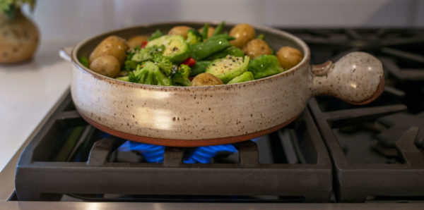 a horizontally framed photograph shows a close up of a clay coyote flameware cazuela filled with cooking veggies and small potatoes. the cazuela is sitting on a lit gas burner, with blue flame visible underneath. the cazuela is glazed in coyote grey (grey with dark speckling through out). the cazuelas main handle is pointed to the right. the vegetables within include: small whole yellow skin on potatoes, broccoli florets, halved brussel sprouts, whole green pea pods and some slices of red bell pepper. course black pepper has been sprinkled onto the cooking vegetables, and they all have a light sheen to them. to the left of the stove top gas range, is a white kitchen countertop. on the countertop is a clay coyote small vase glazed in feather (browns/cream/rust intermingling)