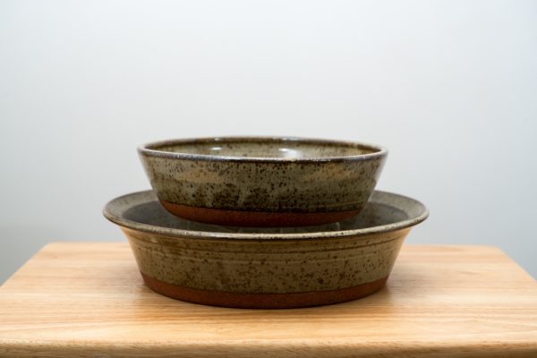 the photograph shows two clay coyote pieces, a savory pie dish with a mini savory pie dish stacked on top of it. the mini savory pie dish is resting on a clear plastic stand allowing it to be almost completely visible white stacked "inside" the regular sized flameware pie dish. both of the pieces are glazed in coyote grey (grey with dark speckling through out, and a unglazed bottom base. the flameware pie dish has larger lip than the mini. both pieces are resting on a small light colored wooden table. the background is a blank white wall. the photograph is lit with white light.