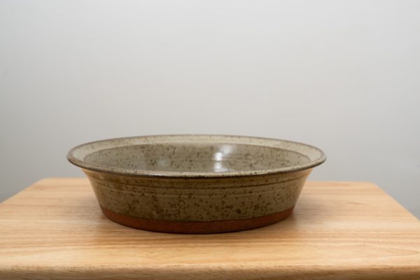 the photograph shows a clay coyote flameware savory pie dish resting on a small light colored wooden table. the flameware savory pie dish is resting flat on the small table. the pie dish is glazed in coyote grey (grey with dark speckling through out). the background is a white wall. the photograph is lit with white light.