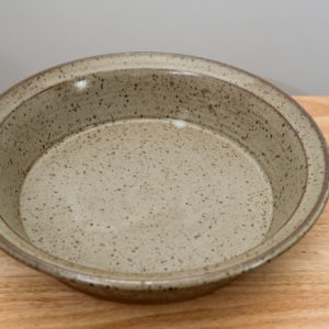 the photograph shows a high angle view of a clay coyote flameware savory pie dish. the angle of the photograph allows the viewer to see the entire cooking surface inside the pie dish. the flameware savory pie dish is glazed in coyote grey (grey with dark speckling through out). the pie dish is resting flat against the small light colored wooden table. the background is a white wall. the photograph is lit with white light.