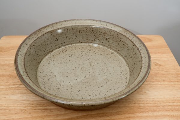 the photograph shows a high angle view of a clay coyote flameware savory pie dish. the angle of the photograph allows the viewer to see the entire cooking surface inside the pie dish. the flameware savory pie dish is glazed in coyote grey (grey with dark speckling through out). the pie dish is resting flat against the small light colored wooden table. the background is a white wall. the photograph is lit with white light.