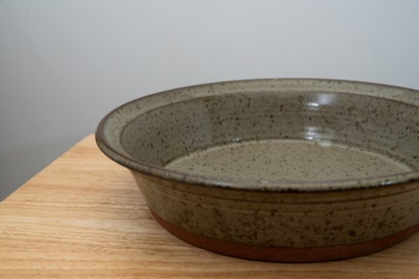 the photograph shows a close up of a clay coyote flameware savory pie dish glazed in coyote grey (grey with darker speckling through out). the flameware savory pie dish is resting flat on the small light colored wooden table. the background is a white wall. the savory pie dish is photographed close enough that it is not 100% in frame, the right side of the flameware savory pie dish is cut out of the picture. the photograph is lit with white light.
