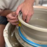 a vertically framed photograph shows a clay coyote potter in process of making a tagine bottom. the photograph is a close up view of a potters hands smoothing/shaping the side of the pottery. the pottery wheel is partially visible. there is a blue plastic circular tray between the wheel surface and the pottery being shaped. the potters hands are on the left side of the pottery, and their (out of focus) body can be partially seen on the left side of the photograph. the wheel, the potter and the pottery are all only partially visible due to the closeness of the shot.