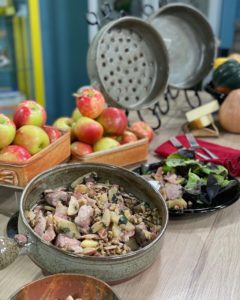 a vertical framed photograph shows a long white wood table filled with clay coyote pottery used to serve and cook a pork, bacon and apple dish. in the foreground is a clay coyote flameware cazuela filled with cooked pork tenderloin, apple slices, herbs and walnuts. behind it is a clay coyote dinner plate glazed in midnight garden, with a serving of the pork dish and a side salad. behind the cazuela with the pork dish, and to the upper left is two clay coyote square bakers glazed in yellow salt, filled with fresh orchard apples, they are bright red and green. further down the table a red napkin with two forks is next to the dinner plate with pork and salad. there is also a clay coyote flameware grill basket and cazuela next to each other, propped up on wire show racks, so they can be easily seen. in front of these two flameware pieces is a clay coyote little dipper, with two sticks of butter resting on it. the little dipper is glazed in yellow salt partially out of frame on the right side of the photo to the viewers perspective are some squashes. the background is out of focus but has blue walls with some bright yellow trim in areas.