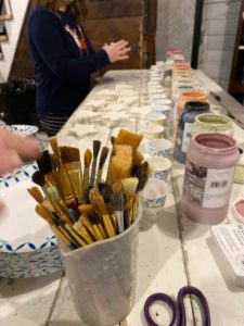 clay coyote holiday ornament making at arts place december 4th 2022 a horizontally framed photograph shows a close up of paint brushes in a small bucket, bristles up. on the right side of the photograph is 3 open plastic jars of colored glaze. between the paint brushes and the plastic jars there is a line small paper cups going parallel to the glaze jars. at the mid top center of the photograph there are stacks of ornaments on the table. on the very left of the photograph, blurred due to motion is a hand reaching for a paint brush.