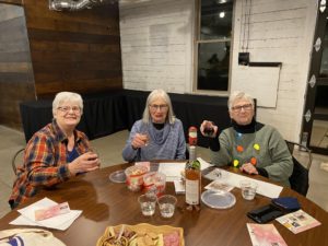 clay coyote holiday ornament making at arts place december 4th 2022 a horizontally framed photograph shows three adults sitting at a circular wooden table. they are all holding up a glass of wine in their right hands. the women on the far right has a light up christmas bulb necklace, and a large sweater. the women in the center has a blue and white striped turtleneck sweater. the women on the left has a burnt orange and navy blue flannel patterned shirt. in the background there is a window, showing it is dark outside. there is a " L " shaped table covered in a long black tablecloth, which reaches to the floor. the wall is white wooden boards, changing to dark wood planks to the left. the floor appears to be cement. on the table in front of the 3 people is snack plates, two small containers shaped like buckets ,but just a bit larger then cups filled with a popcorn mixture. there are two bottles of wine on the table.