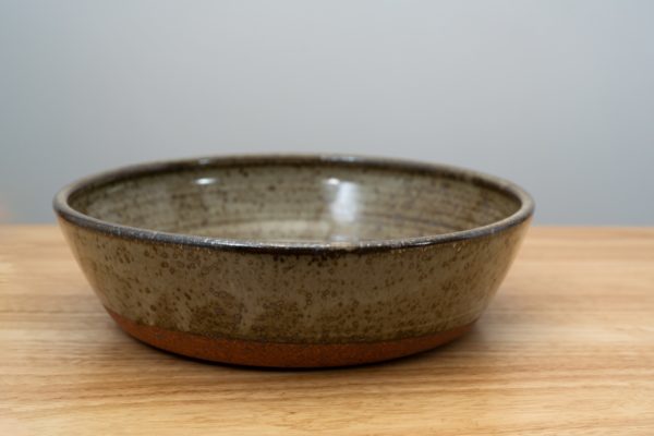 the photograph shows a clay coyote flameware mini savory pie dish resting on a light colored wooden surface. the mini savory pie dish is glazed in coyote grey (grey with dark speckling through out). the min savory is resting flat on the wooden surface. the background is a white wall. the photograph is lit with white light.