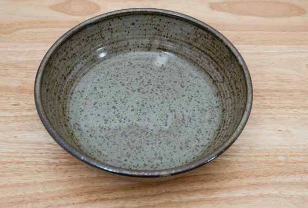 the photograph shows a high angle view of a clay coyote flameware mini savory pie dish resting on a light colored wooden surface. the angle of the photograph allows the entire cooking surface of the mini pie dish to be seen. the flameware mini savory pie dish is glazed in coyote grey (grey with darker speckling through out). the photograph is lit with white light.