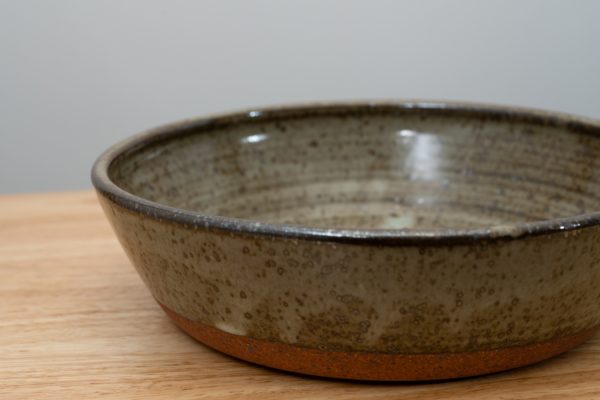 the photograph shows a close up view of a clay coyote flameware mini savory pie dish. the photograph is taken from such a close proximity that the right side and part of the bottom of the flameware mini savory pie dish is cut out of frame. the min savory pie dish is glazed in coyote grey (grey with dark speckling through out). the pie dish is resting on a light colored wooden surface. the background is a plain white wall. the photograph is lit with white light.